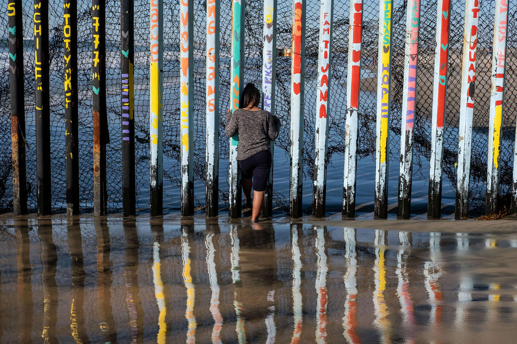 A young girl is alone on the beach and look through the graffiti-covered border fence.