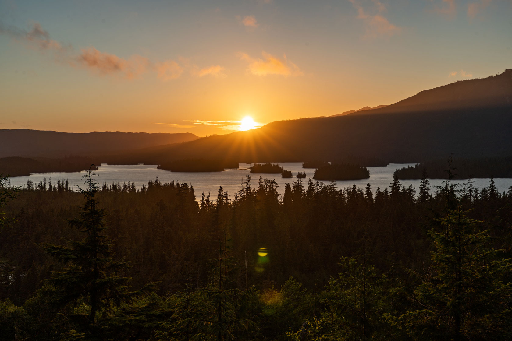 Image showing a sunset in Tongass National Forest, with the sun setting behind some hills in the background, and pine trees populating small islands in the foreground.