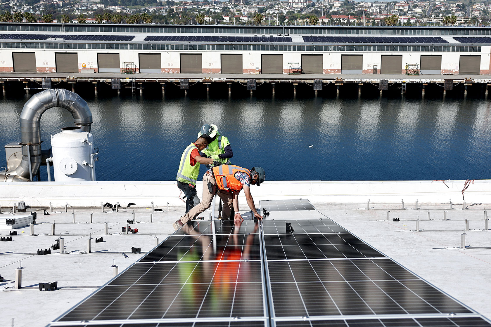 Photo shows two workers wearing bright clothing standing next to a few solar panels on a roof with a river in the background