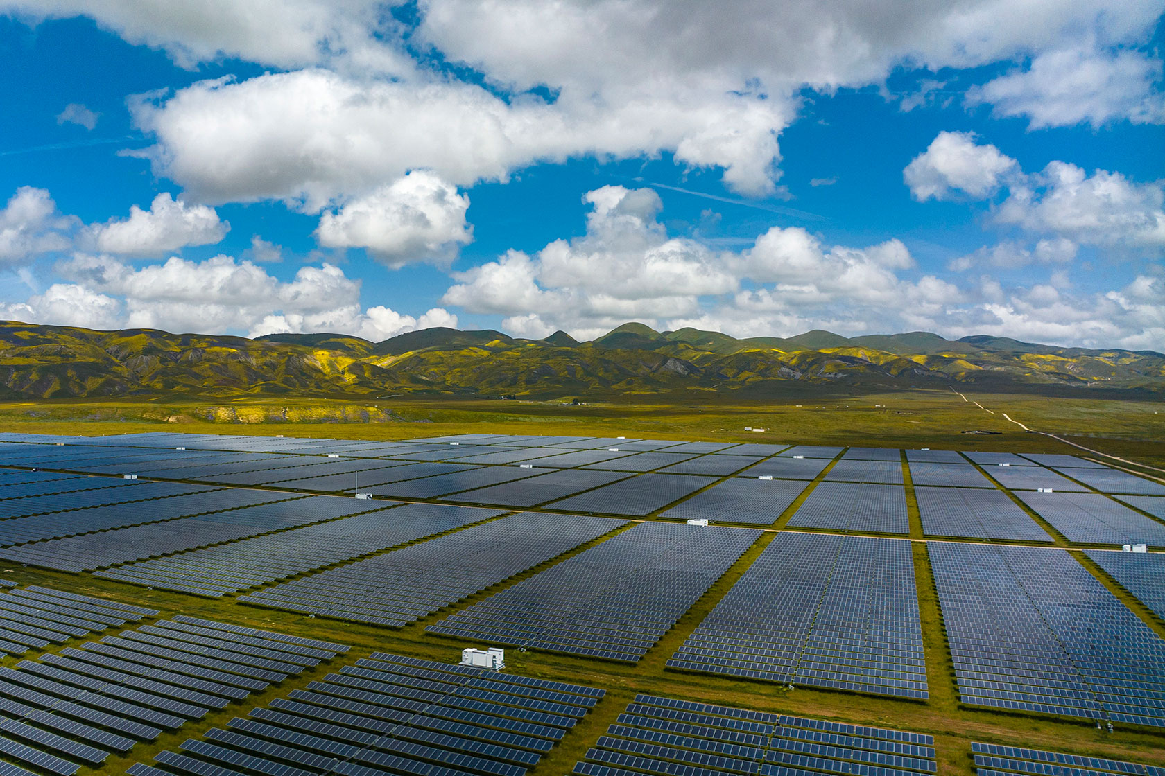 Photo shows an aerial shot of a large solar plant with green hills in the background on a partly cloudy day