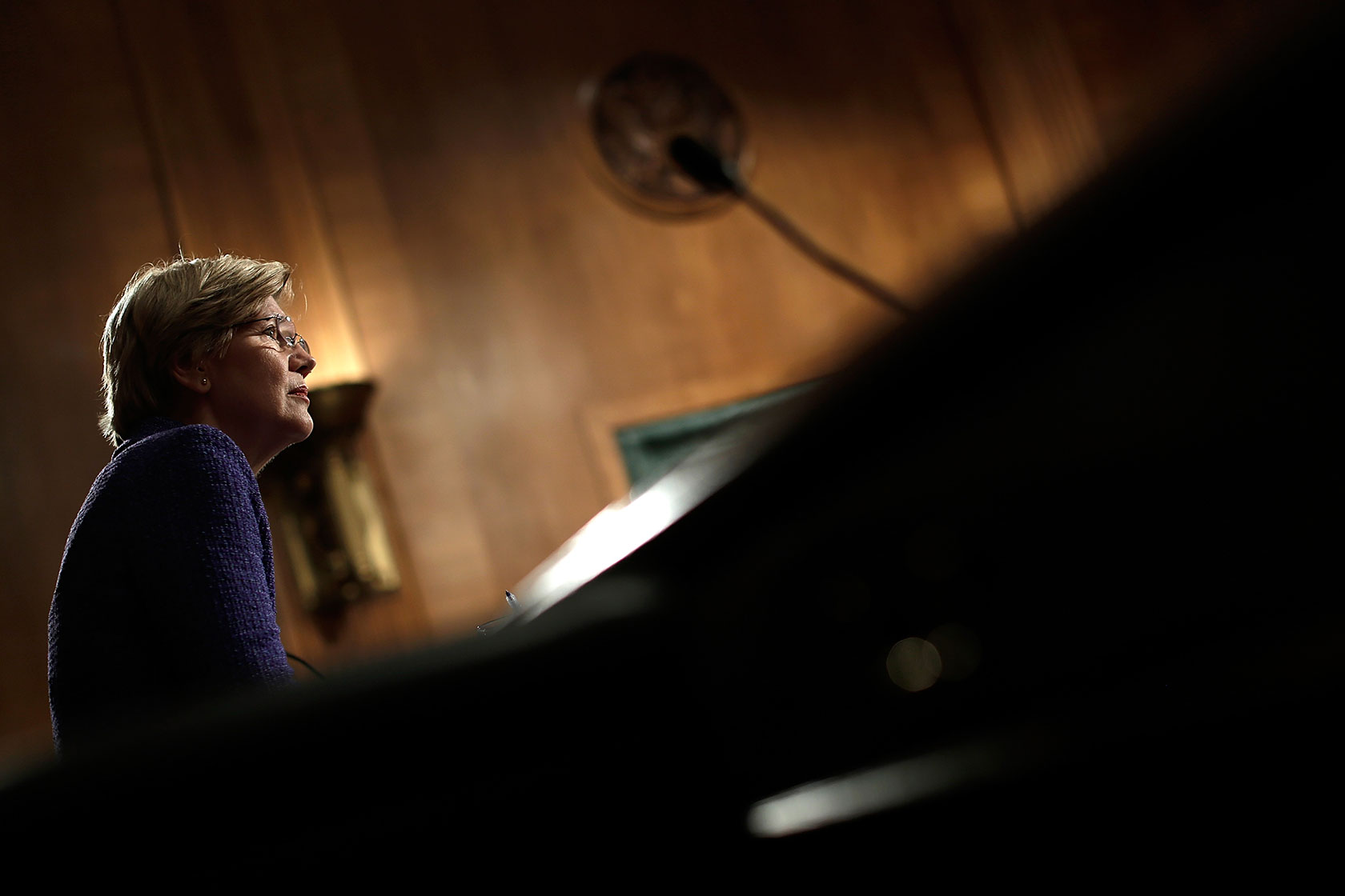 Photo shows Elizabeth Warren backlit in a room with wood paneling