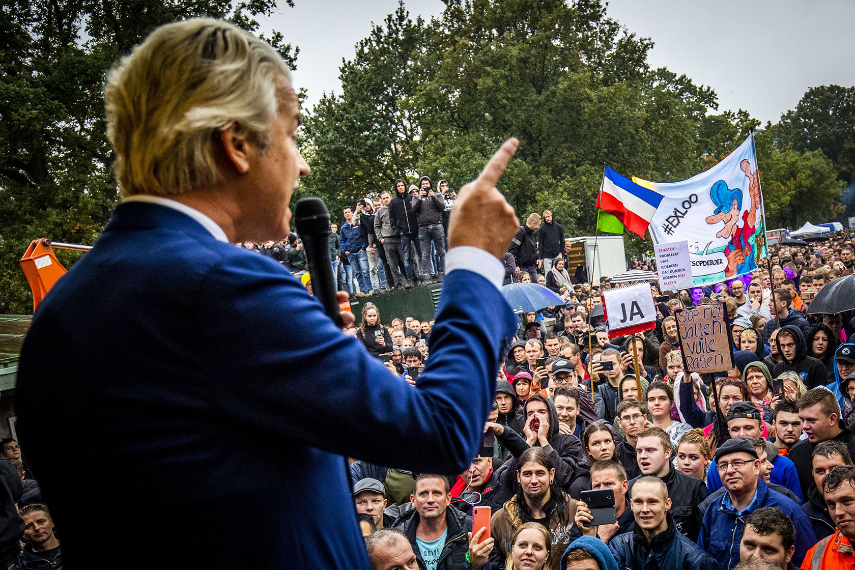 Photo shows the back of Geert Wilders wearing a blue suit in the foreground, slightly blurry, and a large crowd facing him as he speaks, with trees in the background