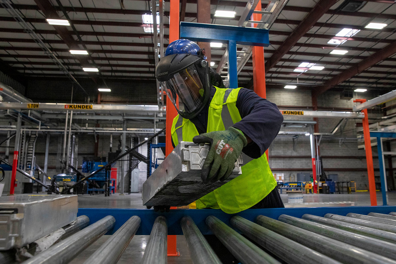 Photo shows a worker in a neon green vest and protective gear placing a battery on a metal conveyor belt with overhead lights in the background