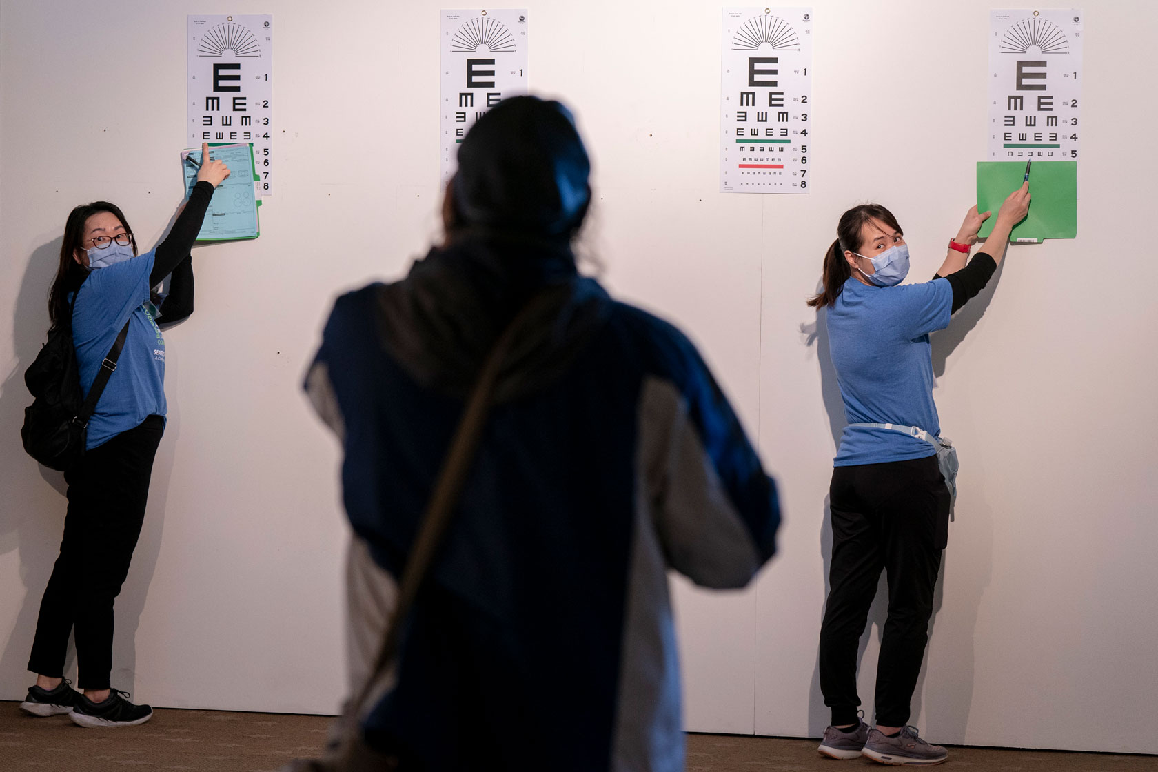 A person stands with their back to the camera in front of two people each pointing to an eye chart.