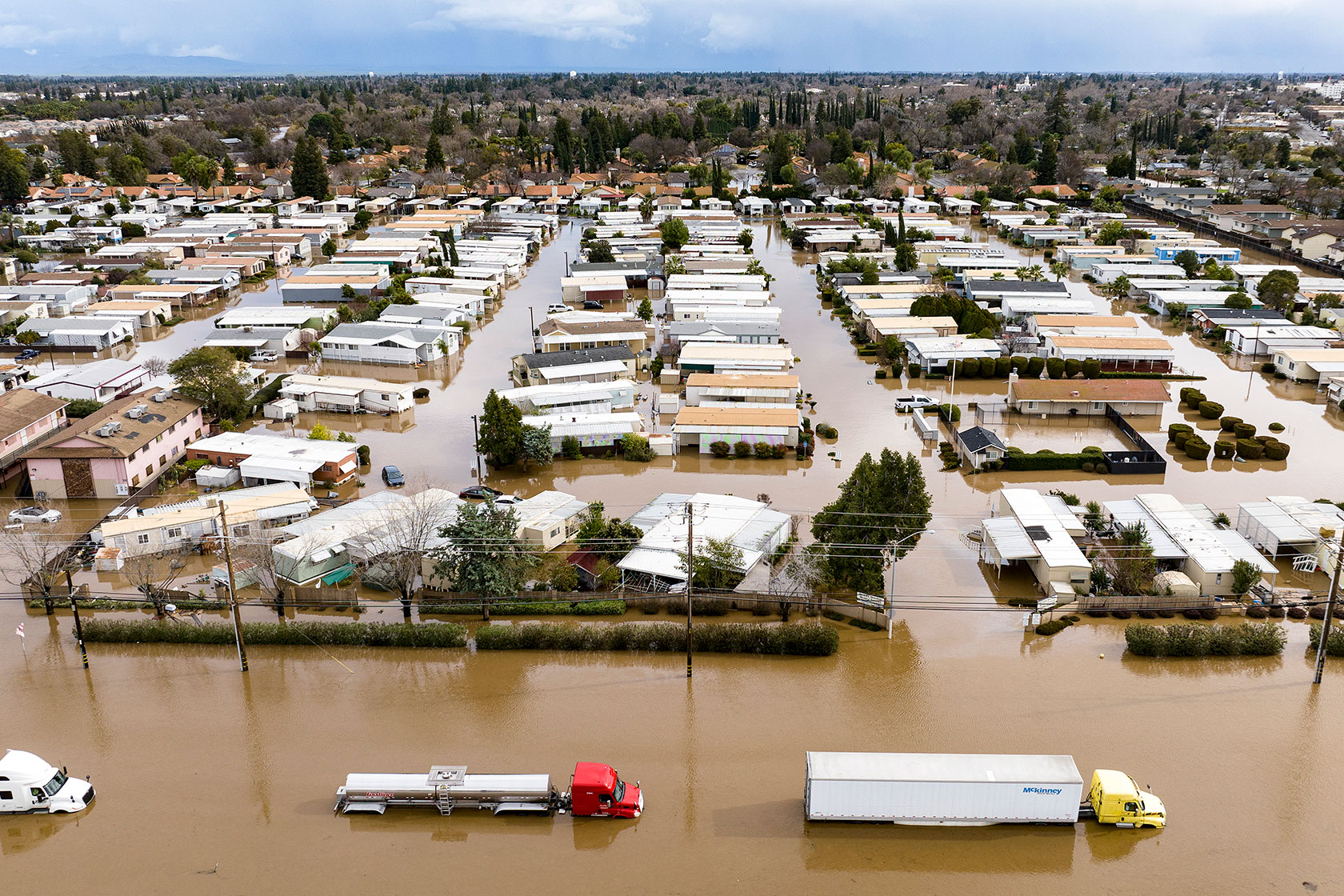 Photo shows brown water flooding streets within a neighborhood against a partly cloudy sky, with homes and trucks partly submerged