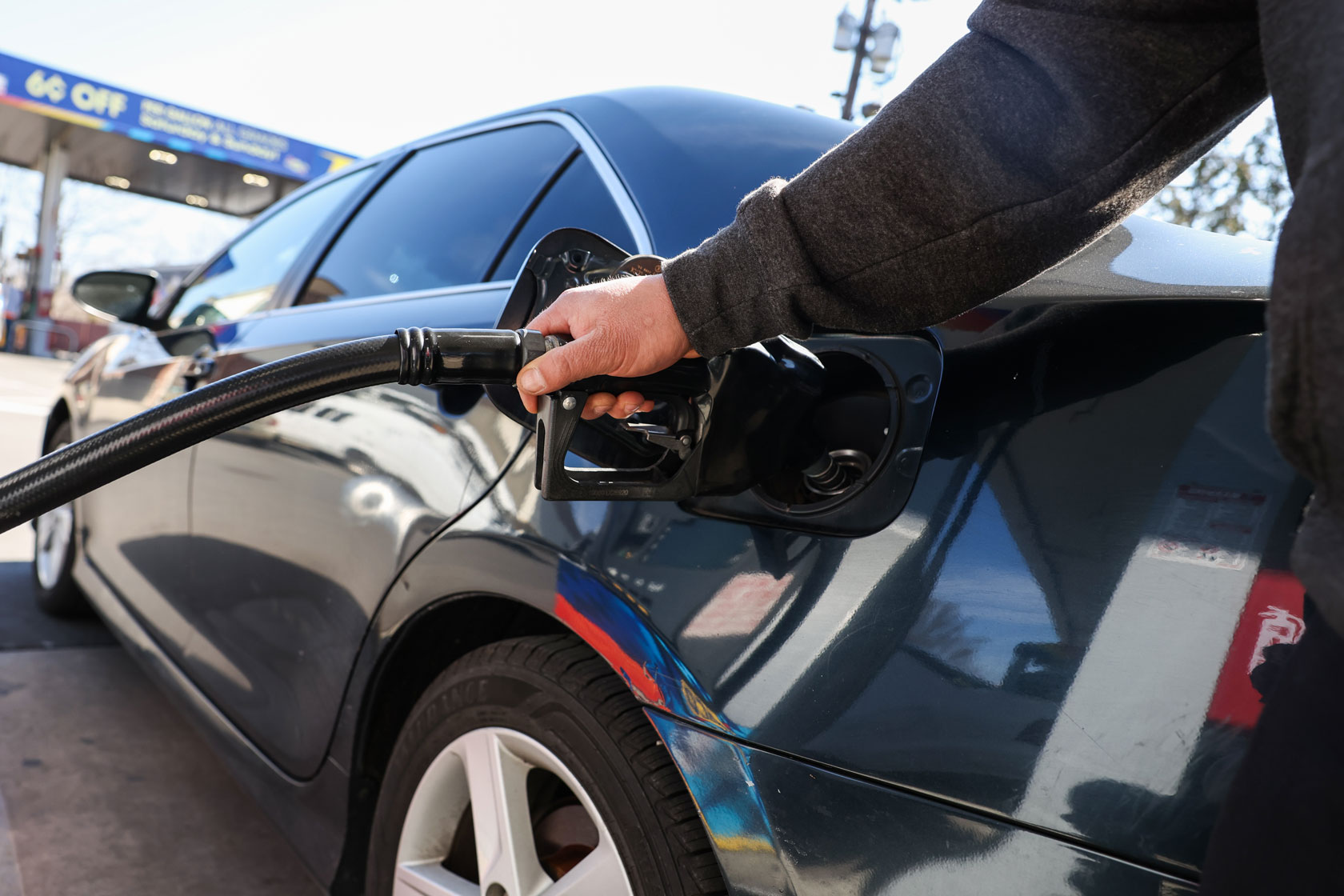 A hand is seen holding a gas pump nozzle up to the gas tank of a black sedan.