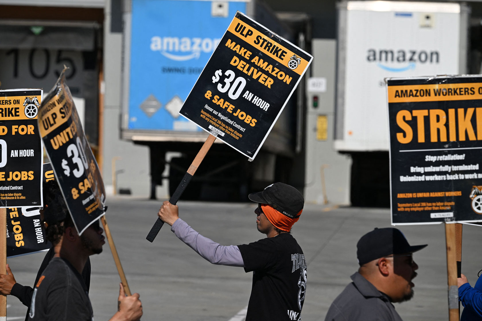 A line of workers holding picket signs marches outside an Amazon warehouse with two delivery trucks parked at it.