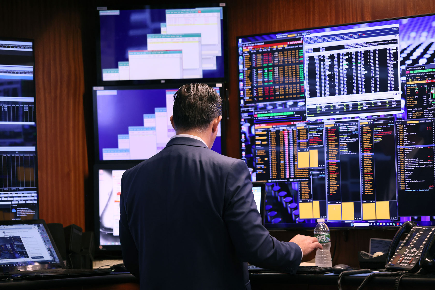 A man stands in front of multiple computer screens displaying stocks.