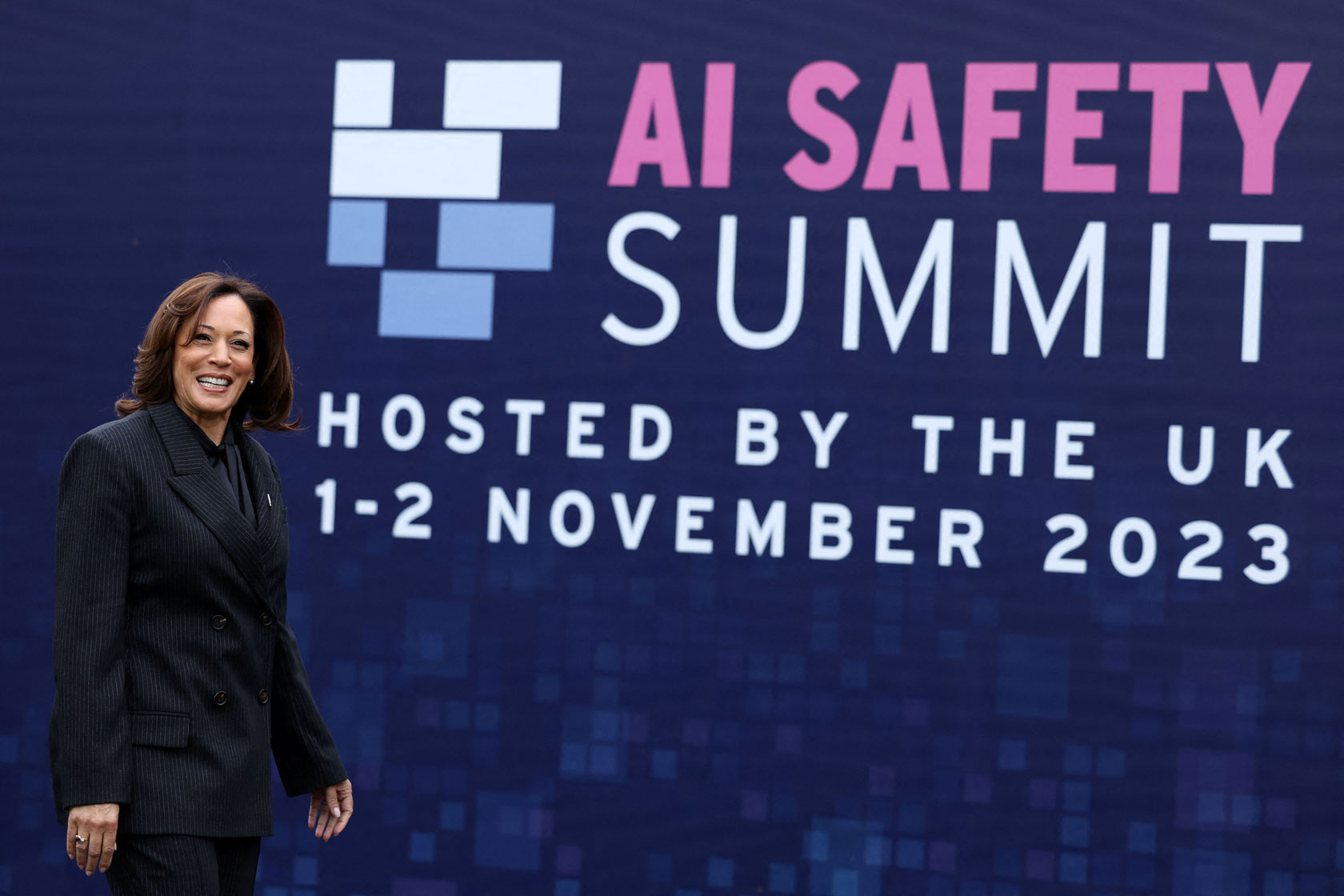 U.S. Vice President Kamala Harris stands smiling to the left of a sign for the AI Safety Summit.