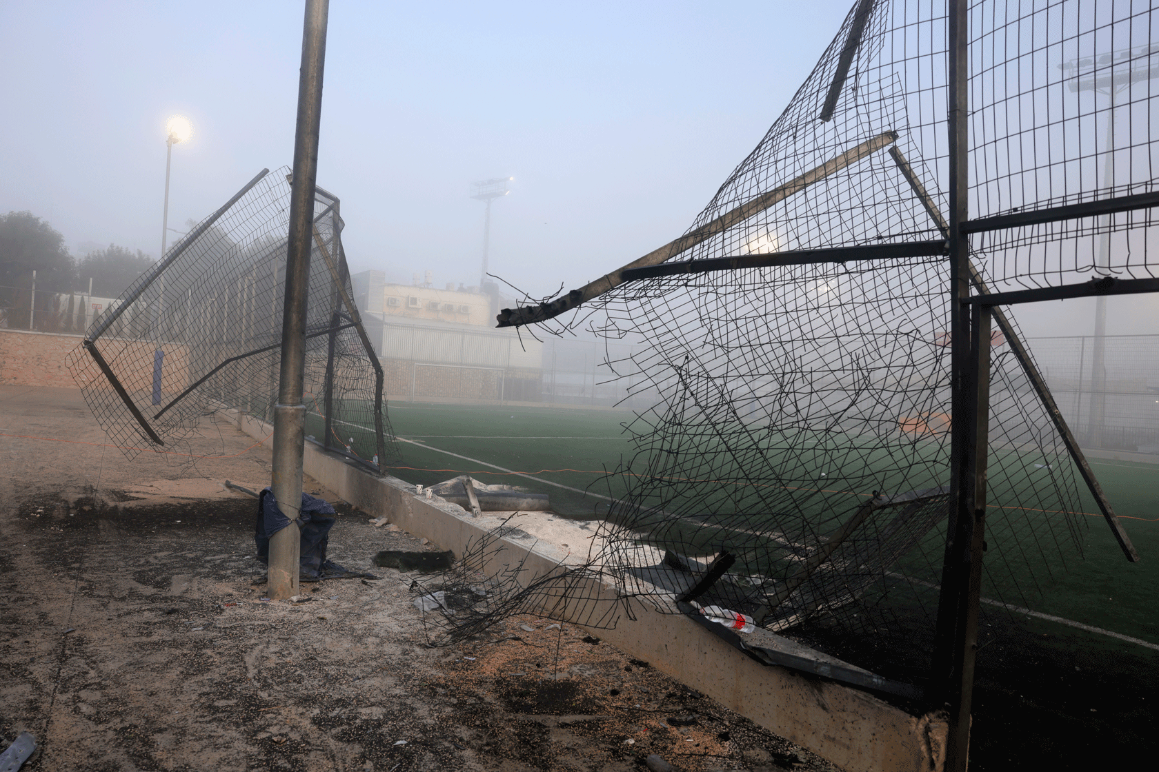 A damaged gate is seen with a socer field in the background.